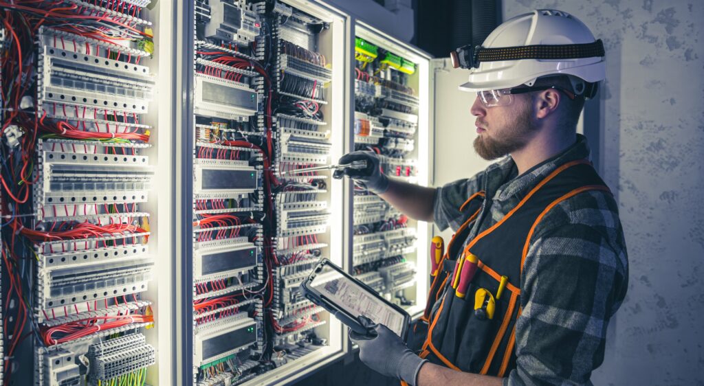Engineer in protective equipment taking readings at an industrial electrical panel with a tablet