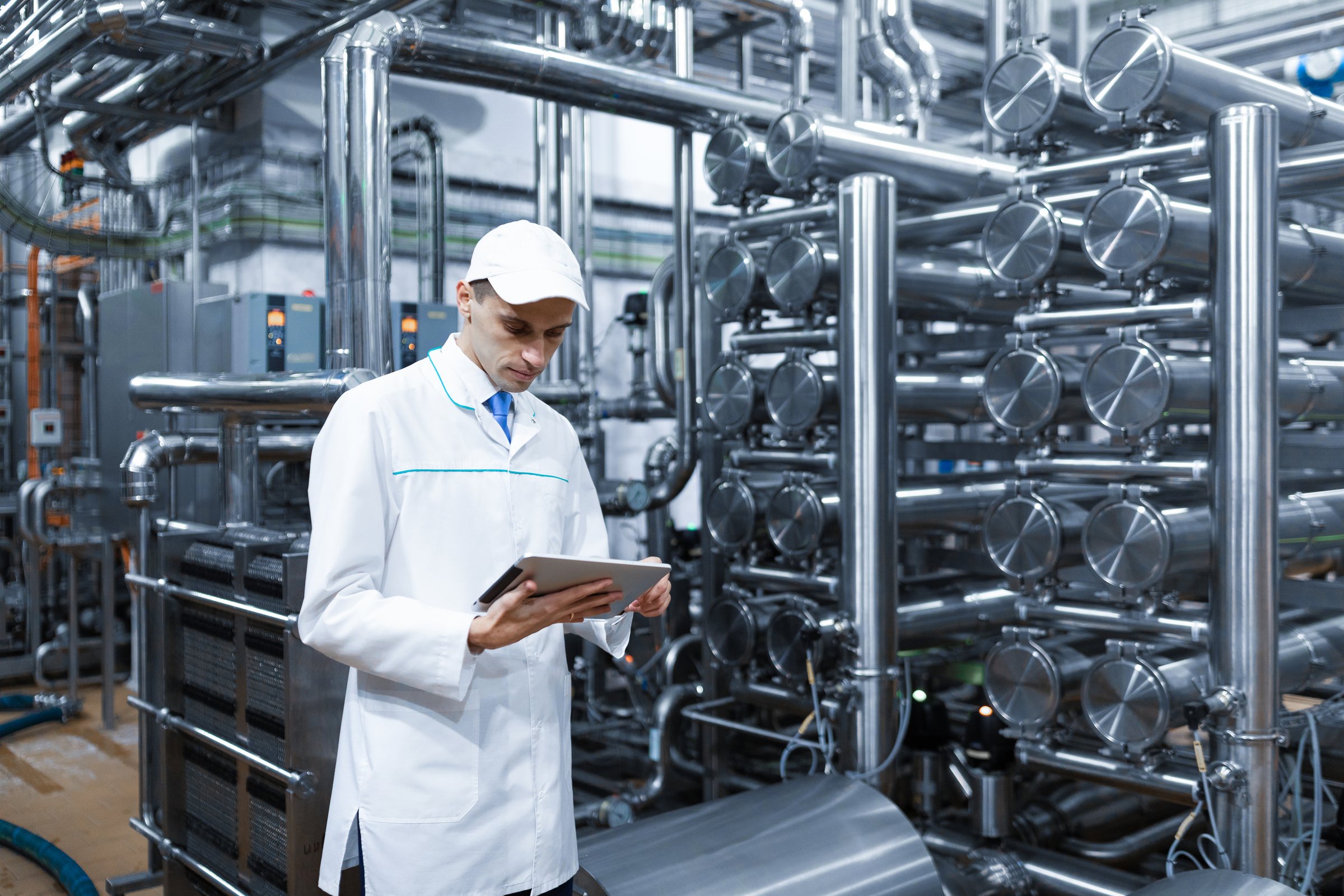 A process engineer in a white coat reviews plant data on a tablet beside stainless steel pipework and vessels